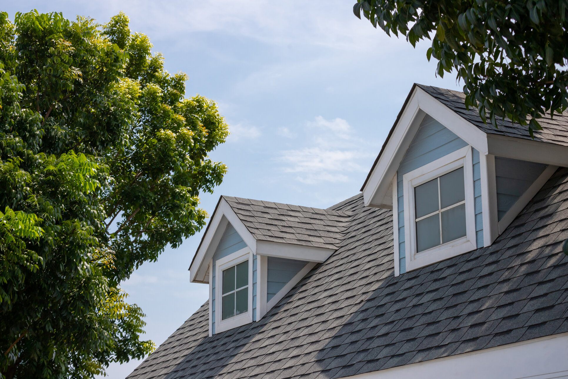 Close-up of a house roof with dark asphalt shingles and two dormer windows framed in white, surrounded by green trees under a clear sky.