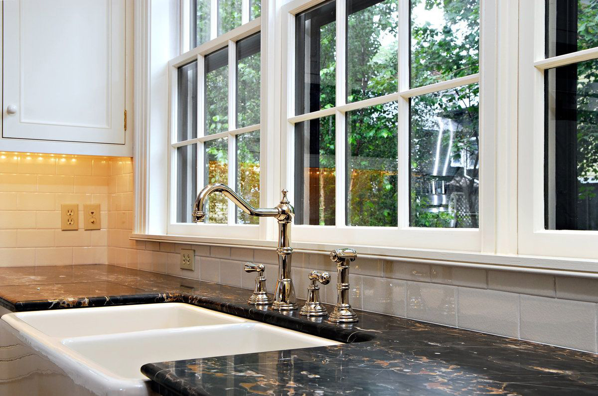 A kitchen interior showing a double white farmhouse sink set into a dark, mottled granite countertop. A shiny chrome gooseneck faucet with matching double handles is centered in front of a large white fiberglass window with muntins, offering a view of green trees outside. White subway tile backsplash and upper cabinets are visible.