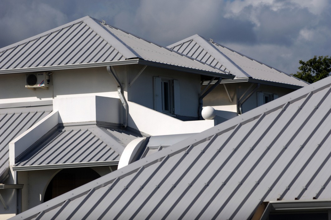 Alt text: Close-up of a gray metal roof with a hip roof design, white siding, and white window trims under a cloudy sky.
