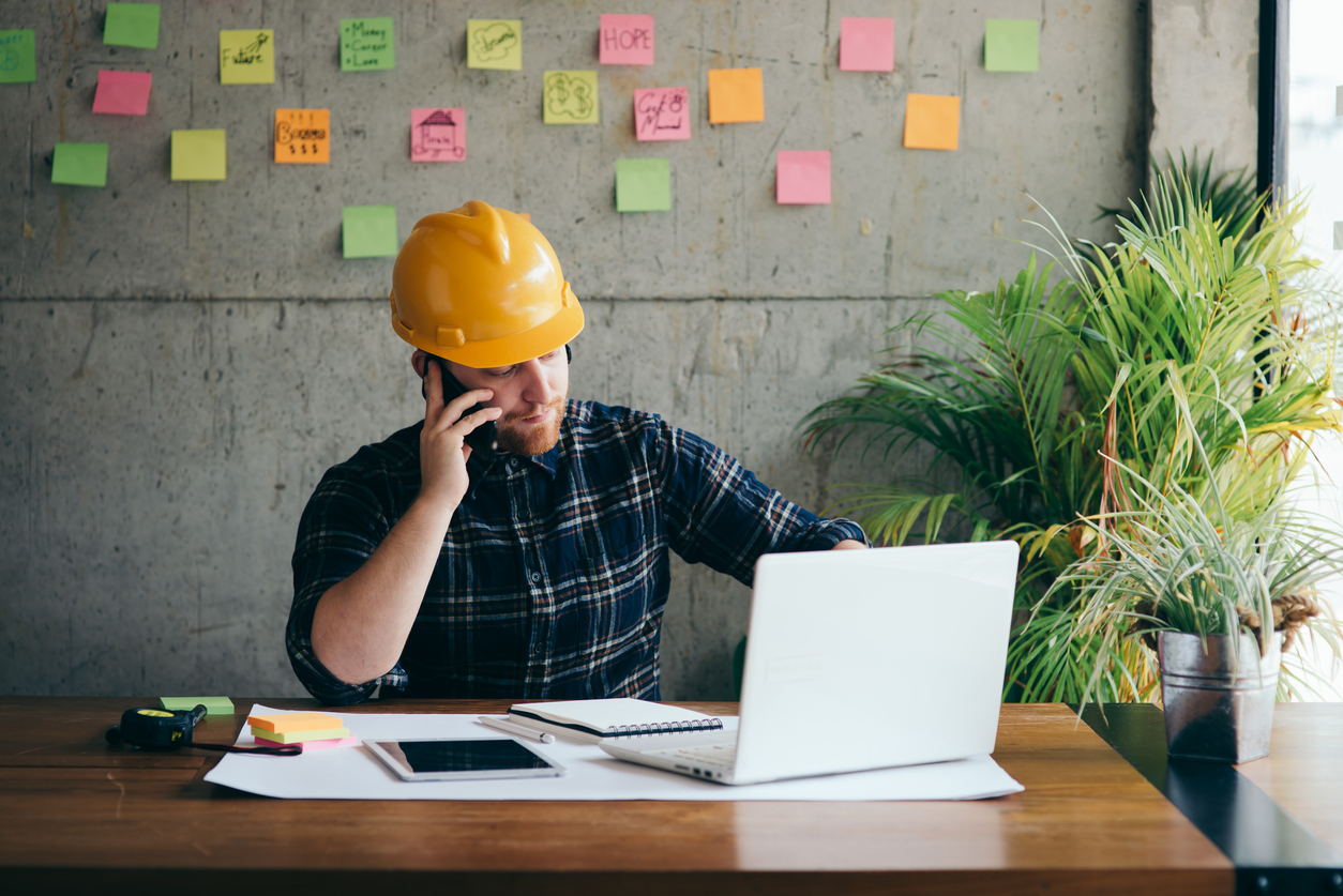 A construction worker in a yellow hard hat and a plaid shirt is sitting at a wooden desk. He is talking on a cell phone while looking at a laptop.