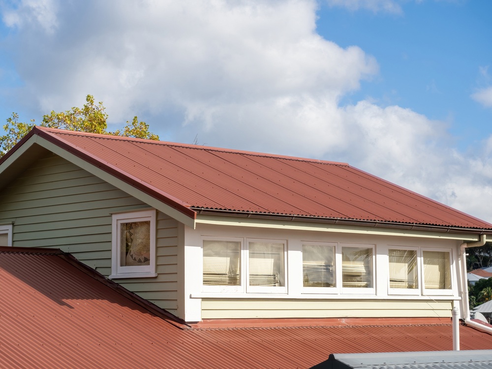 House with a red metal gable roof, light green siding, and white window trims under a partly cloudy blue sky.