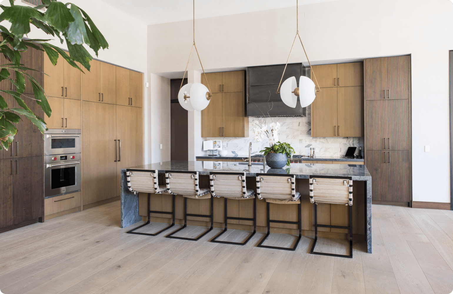 A modern kitchen with a large marble island counter in the center. The island has four wooden chairs with black leather seats around it. There is a stainless steel sink and stovetop on the island counter. In the background, there are light wood cabinets with brass handles and black countertops.