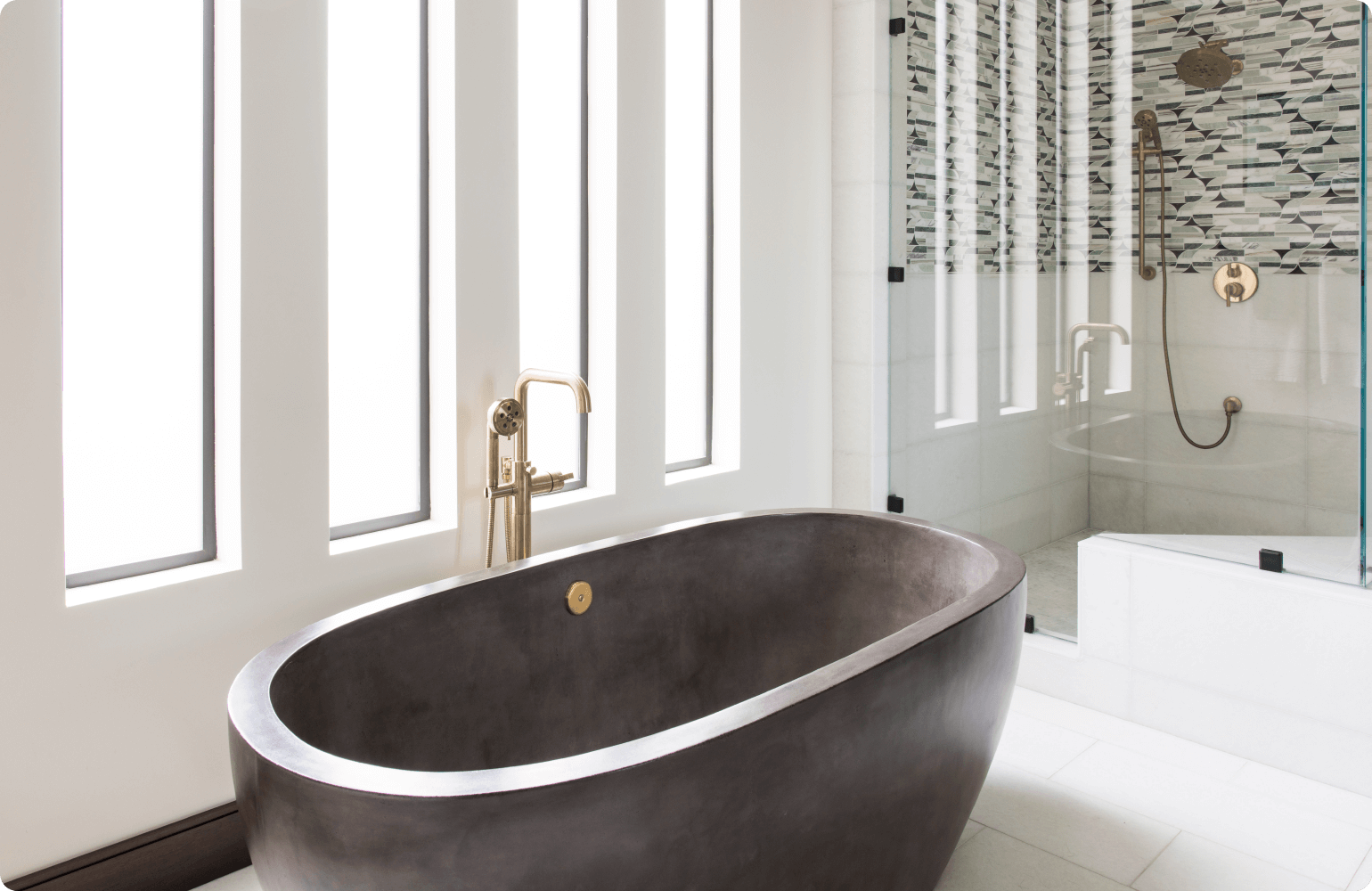 A modern bathroom with a white bathtub and a glass-enclosed shower. The floor and shower walls are tiled in gray marble. There is a single sink vanity with a white countertop and a chrome faucet. The mirror above the vanity is rectangular and has a chrome frame. The toilet is located to the right of the vanity. There is a window on the left wall of the bathroom, which lets in natural light.